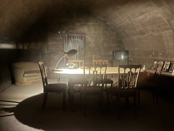 Ricostruzione di una sala riunioni sotterranea con tavolo in legno, sedie d'epoca, lampada ministeriale e ritratti storici.