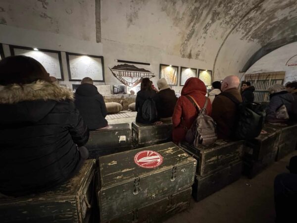 Gruppo di visitatori seduti su casse di legno ascoltano la guida all'interno di una galleria del bunker.