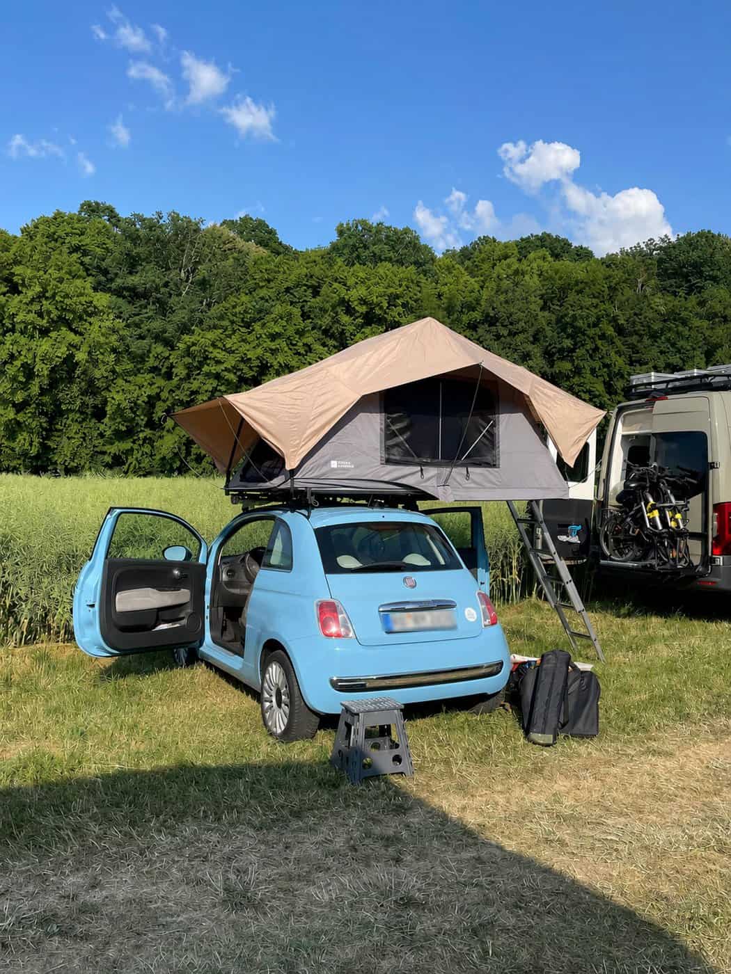 Fiat 500 azzurra con tenda da tetto aperta e scaletta, accanto a un van, ai margini di un campo