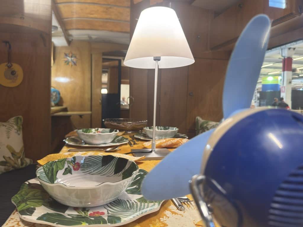 Ventilatore vintage blu in primo piano sul tavolo apparecchiato di una caravan d'epoca La Tortue, con lampada da tavolo e interni in legno, esposta al salone Fit Your Camper