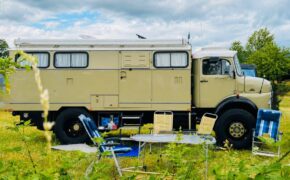 Camper costruito su base Iveco con allestimento minimalista in un prato, circondato da sedie da campeggio e attrezzatura outdoor