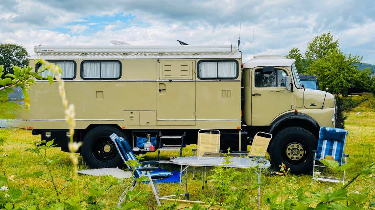 Camper costruito su base Iveco con allestimento minimalista in un prato, circondato da sedie da campeggio e attrezzatura outdoor