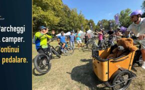 Prova di biciclette a BikeUP al Parco della Cittadella, con cargo bike in primo piano. Testo: Parcheggi il camper. Continui a pedalare.
