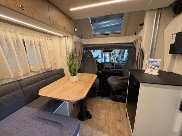 Dinette dell’Atlantis a interni chiari con divano in tessuto grigio, tavolo in legno chiaro, tende e oblò, vista verso la cabina