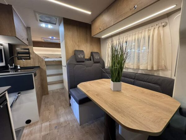 Interno dell’Atlantis a interni chiari: dinette con tavolo in legno, cucina con piano nero a sinistra e in fondo il letto e il bagno