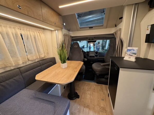 Interno dell’Atlantis a interni chiari: dinette in tessuto grigio, tavolo in legno chiaro, sedili cabina girati e mobile cucina nero