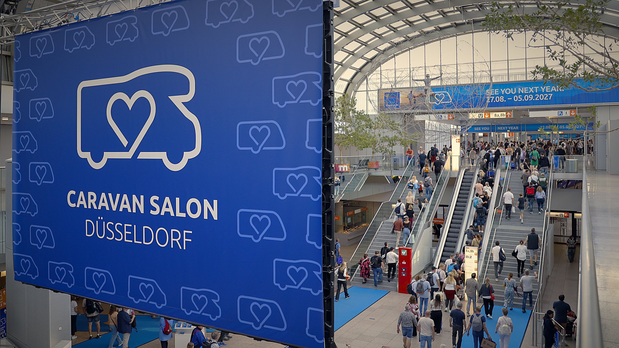 Ingresso del Caravan Salon Düsseldorf 2026 con il banner della fiera e i visitatori sulle scale mobili
